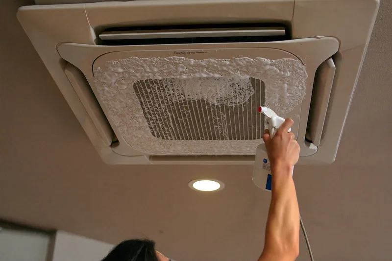 Technician spraying foam cleaner onto the fins of an indoor AC evaporator coil in a villa ceiling
